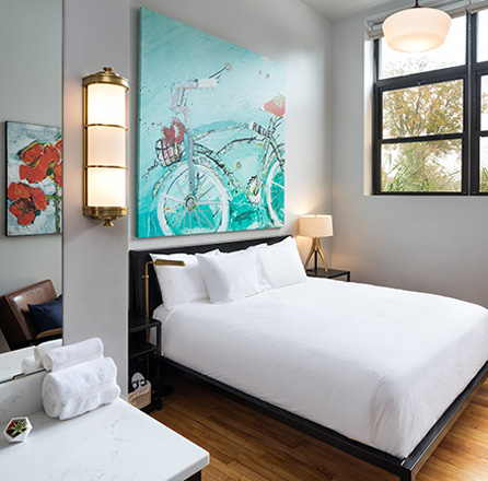 View of a hotel bed room with white sheets and bright paining hanging on the wall behind the bed.